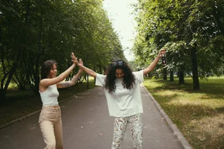 Two women in white attire joyfully dance together in a park, embodying friendship and the spirit of giving.