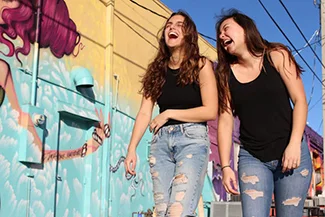 Two young women laugh heartily in front of a colorful mural, embodying the joy of friendship and shared experiences