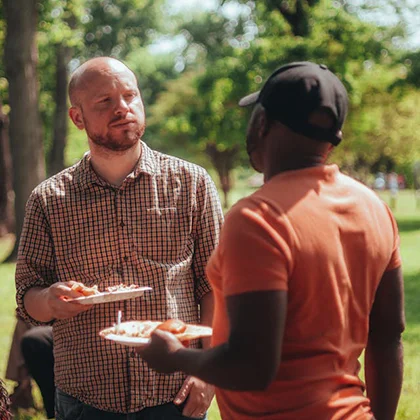 Two man stands in a park, each holding a plate of food, illustrating the simple yet impactful nature of friendship.