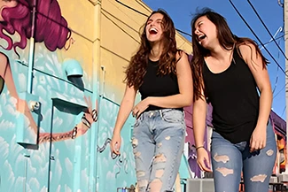 Two young women laugh heartily in front of a colorful mural, embodying the joy of friendship and shared experiences