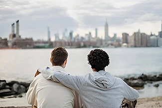 Two friends relax on the beach, a bustling cityscape behind them, illustrating the importance of friendship in a harmonious world.