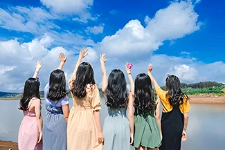 Six girls joyfully stand by a lake, hands raised, celebrating the priceless bond of friendship in a serene setting.