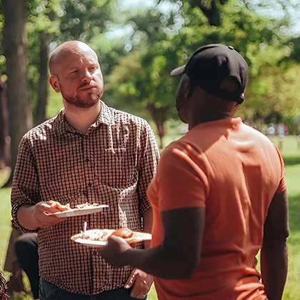 Two man stands in a park, each holding a plate of food, illustrating the simple yet impactful nature of friendship.
