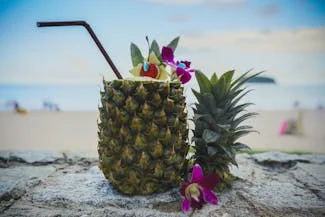 A refreshing pineapple cocktail sits on a beach, surrounded by vibrant tropical flowers under a clear blue sky.