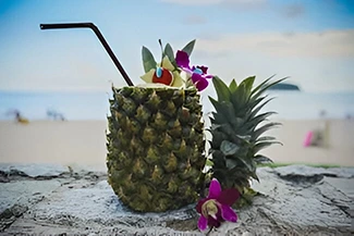 A refreshing pineapple cocktail sits on a beach, surrounded by vibrant tropical flowers under a clear blue sky.