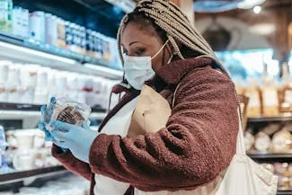 Woman in a grocery store wearing a face mask and gloves, checking food labels for healthier options.