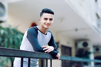 A young man leans on a railing, smiling in front of a building, embodying the message: stress less, smile more.