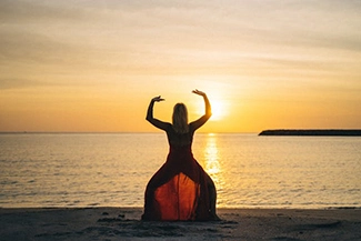 A woman in a flowing red dress stands on the beach at sunset, symbolizing serenity and the joy of movement in nature.