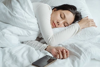 A woman comfortably sleeping in bed, her phone close by, illustrating that sleep is a vital necessity, not a luxury.