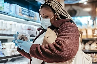 Woman in a grocery store wearing a face mask and gloves, checking food labels for healthier options.