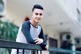 A young man leans on a railing, smiling in front of a building, embodying the message: stress less, smile more.