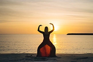 A woman in a flowing red dress stands on the beach at sunset, symbolizing serenity and the joy of movement in nature.
