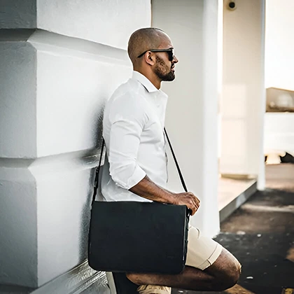 A man in a white shirt and sunglasses leans against a wall, holding a black leather bag, embodying confidence and opportunity.