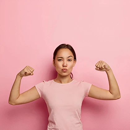 A woman proudly flexes her muscles in front of a pink backdrop, representing strength and the potential for greatness.
