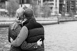 In a serene black and white scene, a couple shares a warm hug by the water, embodying appreciation and connection.