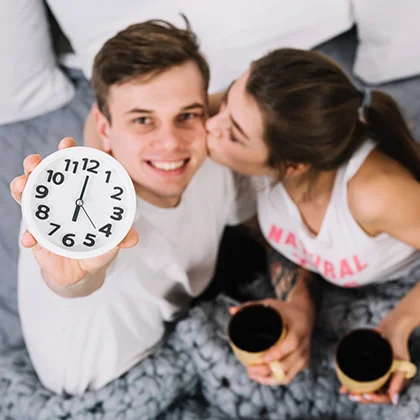 A man and woman relax in bed together, a clock nearby, symbolizing the importance of making time for one another.