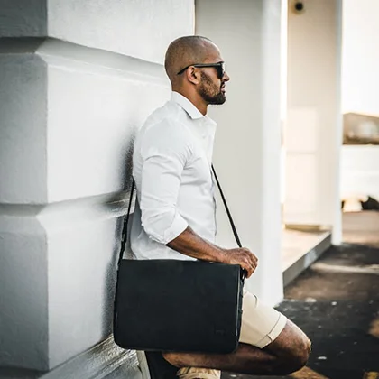 A man in a white shirt and sunglasses leans against a wall, holding a black leather bag, embodying confidence and opportunity.