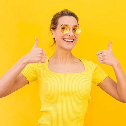 A woman in a yellow shirt and sunglasses smiles while giving a thumbs up, embodying confidence and self-belief.