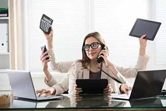 A woman sits at her desk with a laptop and phone, showcasing multitasking skills.