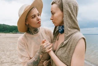 Two women with tattoos on their arms stand together on the beach, embodying empathy and understanding in their presence.