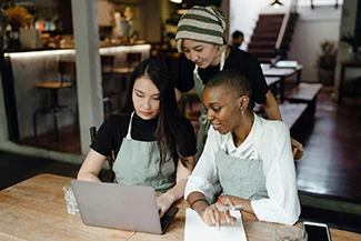 Three women collaborate on a laptop in a restaurant, symbolizing the rise of female entrepreneurship and innovation.