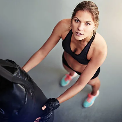 A determined woman in a sports bra and shorts stands confidently beside a punching bag, embodying strength and resilience.