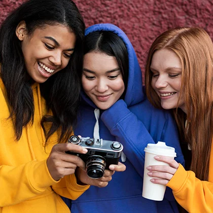 Three girls wearing yellow hoodies pose with a camera, representing women's cultural impact through art and media.